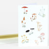 A folded greeting card featuring multiple small watercolor-style mushroom illustrations in red, orange, yellow, black, cream, and pink.