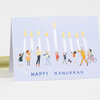 A folded greeting card with an illustrated row of people holding tall lit candles and the text "HAPPY HANUKKAH" on the front.