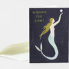 A folded greeting card with a dark cover showing the text "WISHING YOU LIGHT" and an illustrated mermaid reaching toward a small moon or orb.