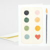 A folded greeting card with an abstract paint-palette design, showing six colored circular swatches and a red heart-shaped swatch beside a vertical paintbrush illustration.