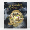 A hardcover book titled "A Garden of Creatures" with an illustrated cover showing a round tan creature holding flowers, a small yellow bird, and a white rabbit, with the names "SHEILA HETI" and "pictures by ESME SHAPIRO" at the bottom.