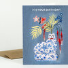 A greeting card with the text “IT’S YOUR BIRTHDAY!” and an illustration of a blue-and-white patterned cat with a red bow sitting under colorful flowers and leaves.