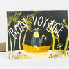A folded greeting card shows the words "BON VOYAGE" over an illustration of a person in a yellow boat among stylized palm trees and foliage on a dark background.