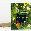 A greeting card with a black cat face hidden in green foliage and flowers, with the text "Happy Birthday!" on the front.