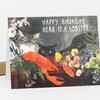 A folded greeting card features an illustration of a bright orange lobster surrounded by flowers, fruit, a cat, and a dog, with the text "HAPPY BIRTHDAY, HERE IS A LOBSTER."