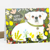 A folded greeting card featuring a white puppy illustration surrounded by colorful flowers and foliage with the text "HAPPY BIRTHDAY!" on the front.