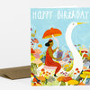 A folded greeting card with the words "HAPPY BIRTHDAY" at the top and an illustration of a woman in red riding a white swan among colorful mushrooms and plants.