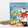 A folded greeting card with the text "HAPPY HANUKKAH!" and an illustration of two white cats, a menorah, and holiday food.