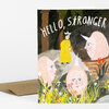 A folded greeting card with the text "HELLO, STRANGER" and an illustrated scene of a person in a yellow coat standing among three pink, horned figures and flowers.