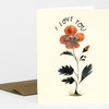 A folded greeting card with a cream front featuring a flower illustration with a face and the words "I LOVE YOU" arched above it.