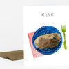 A folded greeting card with the text "PRE LATKE" above an illustration of a blue plate holding a latke, with a red striped napkin and a green fork beside it.