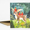 A folded greeting card with the text “TIS the SEASON” above an illustration of three red-clad figures riding a deer in a forest scene.