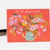 A folded greeting card with an orange front printed with the text "TO THE newlyweds!" above a blue floral watering can holding an arrangement of pink and white flowers and green leaves.