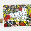 A greeting card front that says “WELCOME, TINY ONE!” and shows a white tiger surrounded by colorful flowers and leaves, with a small child sitting on its head.