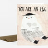 A folded greeting card with the text "YOU ARE AN EGG" above an illustration of a stylized egg-shaped figure wearing a black hat, plaid clothing, and black shoes.
