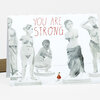 A folded greeting card printed with the text "YOU ARE STRONG." above grayscale drawings of several classical-style nude statues and a tiny red-dressed figure near the bottom center.