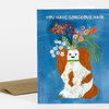 A folded greeting card with a blue cover showing a white dog with orange curly ears and flowers growing from its head, with the text "YOU HAVE GORGEOUS HAIR" at the top.