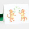 A folded greeting card with two orange cats juggling five green striped ornaments on the front.