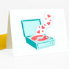 A folded greeting card featuring a teal record player with a red vinyl record and small red hearts rising above it.