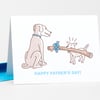 A folded greeting card with a line-drawn dog and small dog carrying a large stick tied with a blue bow, with the text "HAPPY FATHER'S DAY!" on the front.