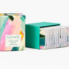 A colorful boxed card game with an abstract painted pattern and a label that reads "FLATTER ME" and "A COMPLIMENT BATTLE CARD GAME," shown with a teal card case and stacked cards.