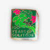 A rectangular enamel pin shaped like a book cover with a green background, gold outline, pink flowers, a small black ant, and the text "100 YEARS OF SOLITUDE".