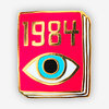 A small enamel pin shaped like a bright pink book cover with gold-outlined edges, the number "1984" at the top, and a stylized eye graphic on the front.