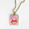 A gold chain necklace with a dangling pink book-shaped charm that reads "Alice in Wonderland" and shows a red grinning cat face on the cover.