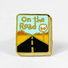 A square enamel pin with gold outline and a stylized highway scene, showing the text "On the Road" and a Route 66 shield.