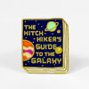 An enamel pin shaped like a book cover with the text "THE HITCH HIKER'S GUIDE TO THE GALAXY," showing a blue space-themed cover with planets and gold-outlined pages.