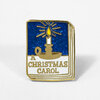 A glittery enamel pin shaped like a book cover with a gold candle icon and the text "A CHRISTMAS CAROL".