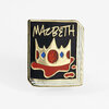 A rectangular enamel pin shaped like a black book cover with gold edges, the word "MACBETH" at the top, and a stylized gold crown with red and blue jewel details over a red drip-like design.