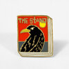 A rectangular enamel pin with gold outline shows a black bird on a red and gray background with the text "THE STAND" at the top and a yellow circle in the upper right.