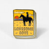 A rectangular enamel pin shaped like a book cover shows a black cowboy on horseback at sunset with the text "LONESOME DOVE".