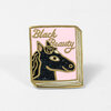 A small enamel pin shaped like a book cover with a pink background, gold outline, glittery black horse head illustration, and the words "Black Beauty."