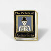 A small enamel pin shaped like a book cover with the text "The Picture of" at the top and "Dorian Gray" at the bottom, featuring a skeleton wearing a top hat in the center.