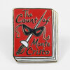 A rectangular enamel pin shaped like a red book cover with gold-toned outlines, showing the text "The Count of Monte Cristo" and a black masquerade mask over a sword.