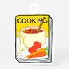 A die-cut sticker shaped like a cookbook cover with the word "COOKING" on the front and an illustration of a soup pot with a spoon, onion, tomatoes, carrots, and garlic cloves.