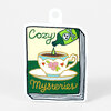 A die-cut sticker shaped like a book cover shows the words "Cozy Mysteries" around an illustration of a teacup with flowers while a green bottle with a skull-and-crossbones label pours a drop into it.