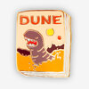 A small enamel pin shaped like a book cover with the word "DUNE" at the top and a stylized illustrated creature on a tan, orange, and brown design.