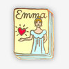 A rectangular enamel pin shaped like a book, with the name "Emma" at the top, a woman in a light blue dress, and a red heart graphic.