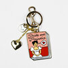 A gold-tone keychain with a book-shaped enamel charm reading "Pride and Prejudice," a small heart charm, and a small face charm attached by a short chain.