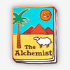 A book-shaped enamel pin shows a colorful cover with a palm tree, desert mountains, a smiling sun, a white sheep, and the text "The Alchemist."
