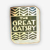 A rectangular enamel-style pin shaped like a book cover with the text "THE GREAT GATSBY" in the center and black, cream, and gold geometric border patterns.