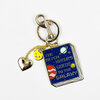 A gold-tone keychain with a book-shaped charm reading “THE HITCH-HIKER'S GUIDE TO THE GALAXY,” plus a small heart charm and a small round yellow smiley-face charm.