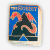 A book-shaped enamel pin with the title "THE HOBBIT" at the top and a red dragon illustration on a blue and black cover design.