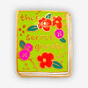 A small rectangular enamel pin shaped like a book, with a bright green cover decorated with flowers and leaves and the text "the secret garden".