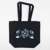 A black tote bag with long handles and a white line drawing of two bugs standing on rocks beside a central floral design.