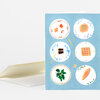 A folded greeting card with a blue front featuring six illustrated plates with different foods and decorative borders, shown with a white envelope.