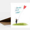A blank white greeting card with the hand-lettered text "FOLLOW YOUR HEART" and an illustration of a small figure holding a string attached to a red heart-shaped balloon above a green hill.